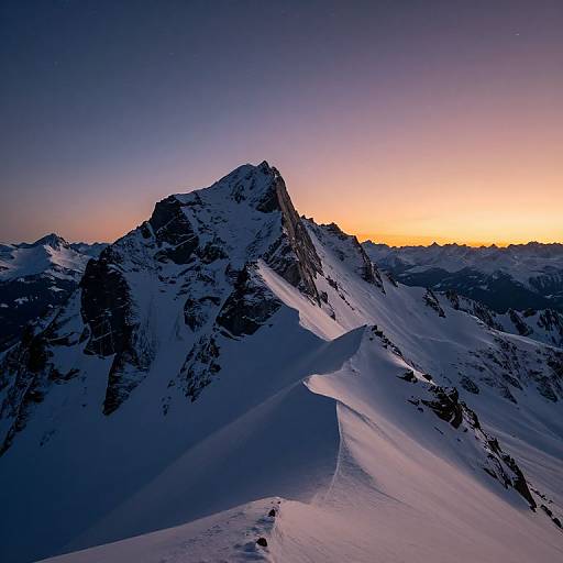 Photograph of a snow-covered mountain peak at sunset, with a gradient sky from deep blue to warm orange, and rugged slopes casting long shadows.