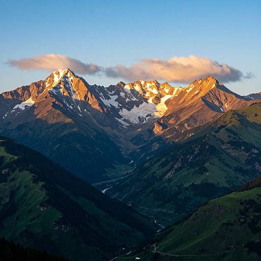 Photograph of a sunlit mountain range with jagged peaks, glowing orange and yellow, under a clear blue sky with a few clouds. Dark green