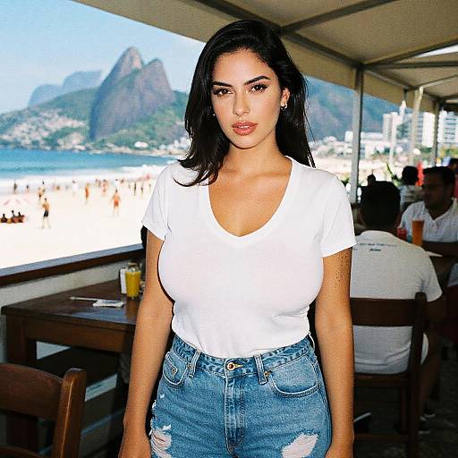 Photograph of a Latina woman with long black hair, wearing a white t-shirt and blue, distressed jeans, standing in a beachside café with mountains