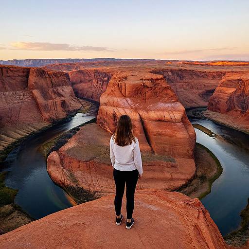 Photograph of a woman with brown hair, white shirt, black pants, and sneakers, standing on a red rock ledge, overlooking a vast, rugged