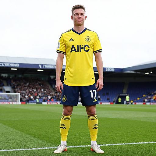 Photograph of a male soccer player standing on a green field, wearing a yellow jersey with 