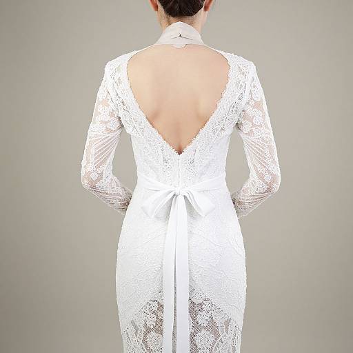 Photograph of a woman from behind, wearing a white lace wedding dress with a deep V-back and long sleeves, against a plain gray background.