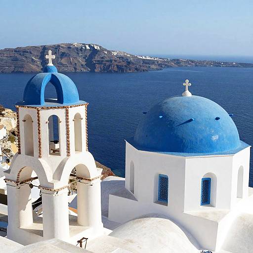 Photograph of two blue-domed Greek Orthodox churches with white walls, set against a calm blue sea and distant rocky hills.