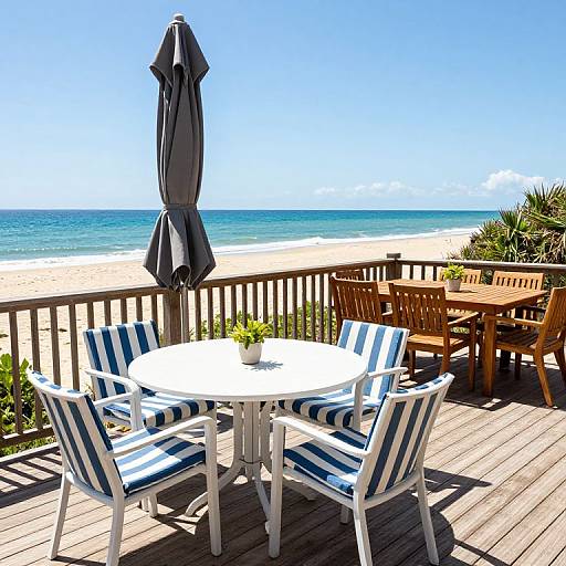 Beachside patio photo: White table with blue-striped chairs, closed gray umbrella, wooden tables, sunny ocean view, clear sky, and sandy beach