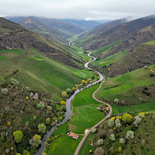 Aerial photograph of a winding river through lush green hills, dotted with small houses, trees, and yellow foliage, under a cloudy sky.