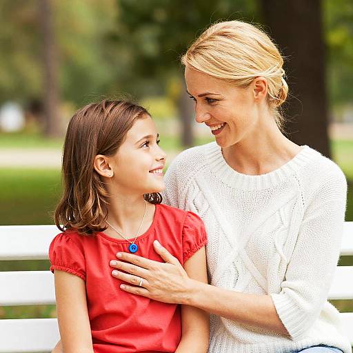 Photograph of a smiling blonde woman in a white knit top gently touching the chest of a brown-haired girl in a red shirt, sitting on a white