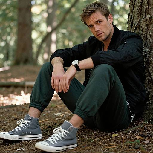 Photograph of a young man with light brown hair and blue eyes, sitting against a tree in a forest, wearing a black jacket, gray pants,