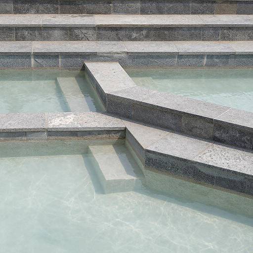 Photograph of a modern, geometric concrete pool design with stepped platforms, casting sharp shadows on clear, turquoise water.