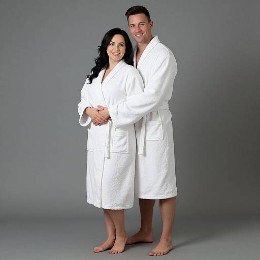 Photograph of a smiling couple standing closely, both wearing white bathrobes, against a plain gray background, with the man behind her.