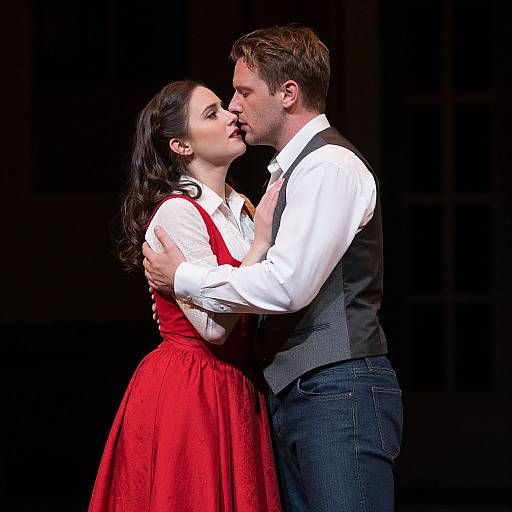 Photograph of a couple passionately kissing on stage; woman in red dress, white blouse, man in white shirt, grey vest, blue jeans. Black