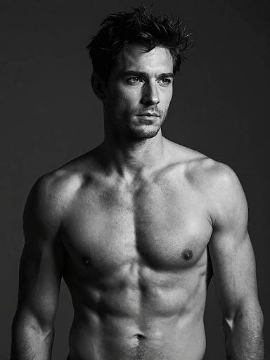 Black-and-white photograph of a muscular, shirtless man with short, dark, tousled hair, standing against a dark gray background.