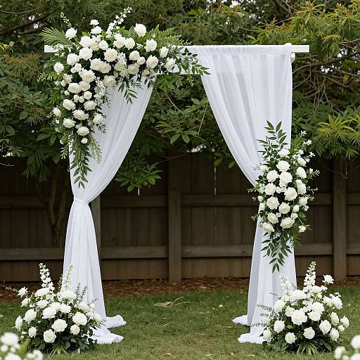 Photograph of a white, floral archway with sheer curtains, adorned with white flowers and greenery, set against a wooden fence and lush greenery