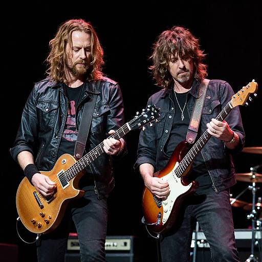 Photograph of two long-haired, bearded male guitarists with messy hair, wearing black leather jackets, playing electric guitars on stage.