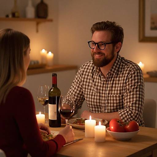 Photograph of a bearded man with glasses, in a checkered shirt, smiling at a woman, candlelit dinner table with wine and tomatoes.
