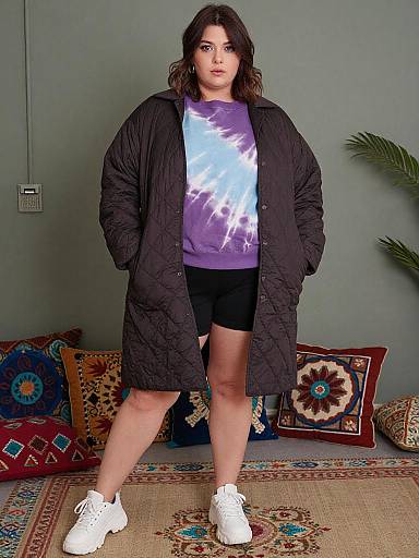 Photograph of a plus-sized woman with dark hair, wearing a black quilted coat, purple graphic tee, black shorts, and white sneakers, standing