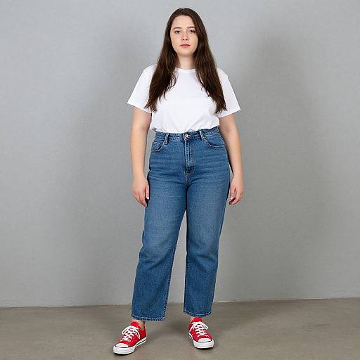 Photograph of a young woman with long dark hair, wearing a white t-shirt, high-waisted blue jeans, and red sneakers, standing against