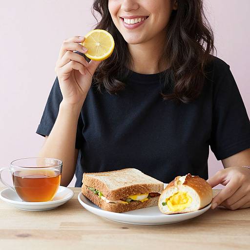 Photograph of a smiling woman with dark hair, black t-shirt, holding a lemon slice over tea, with a sandwich and egg bun on a white