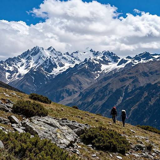 Photograph of two hikers in the foreground on a rocky mountain slope, with snow-capped peaks and a bright blue sky with white clouds in the