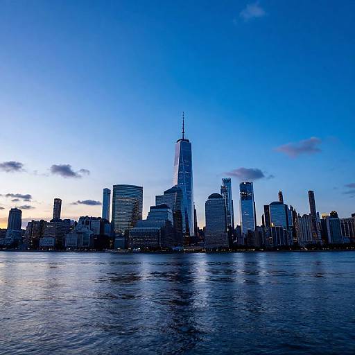 Photograph of New York City's skyline at dusk, featuring the One World Trade Center as the tallest building, with a deep blue sky and calm water