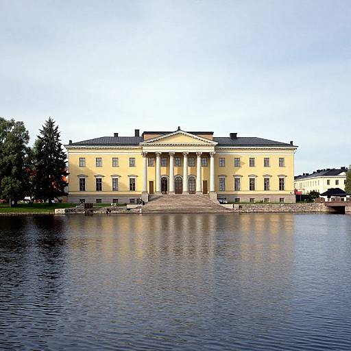 Photograph of a grand, yellow neoclassical building with a central portico, reflecting on a calm river, under a clear blue sky.