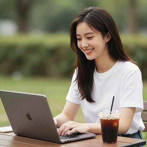 Asian woman with long black hair, smiling, wearing white t-shirt, using Apple laptop outdoors, with iced drink in front. Photographic image.