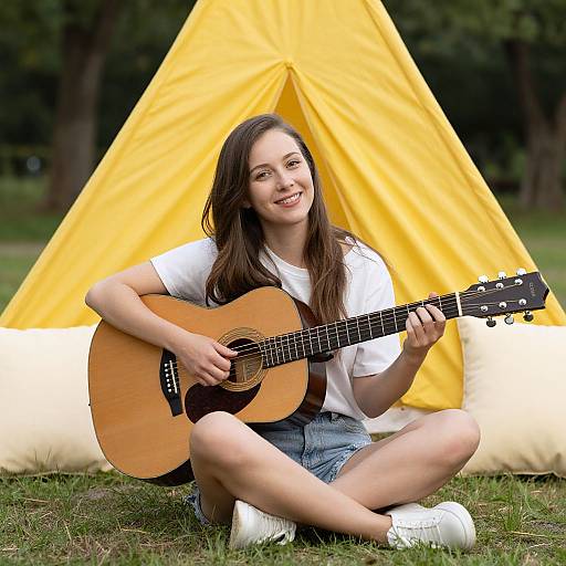 Photograph of a smiling young woman with long brown hair, wearing a white t-shirt and denim shorts, sitting on grass playing an acoustic guitar in front