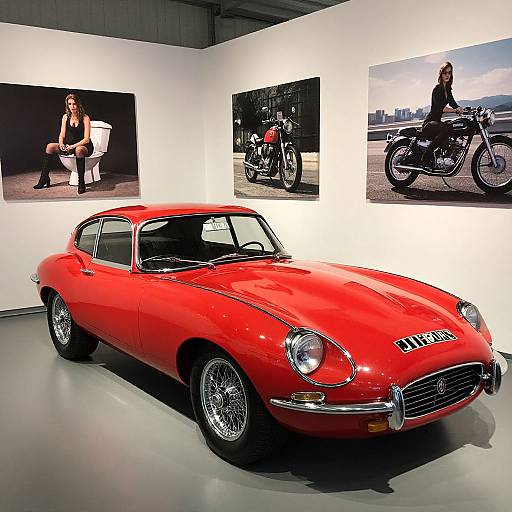 Photograph of a vibrant red vintage sports car in a gallery, flanked by black-and-white motorcycle photos on white walls.