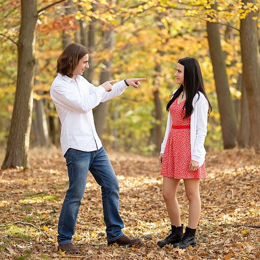 Photograph of a man in a white shirt and blue jeans pointing at a woman in a red polka-dot dress, standing in a fall forest with