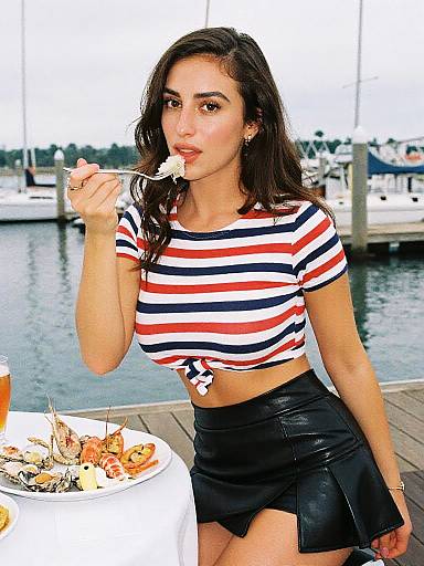 Photograph of a young woman with long dark hair, wearing a striped crop top and black leather skirt, eating seafood by a marina.