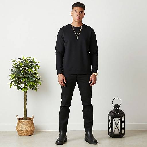 Photograph of a young man with short black hair, wearing a black long-sleeve shirt, pants, and boots, standing against a white wall