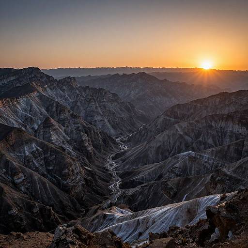 Photograph of a dramatic mountain range at sunset, with the sun low on the horizon casting a golden glow over dark, rugged peaks and a winding river