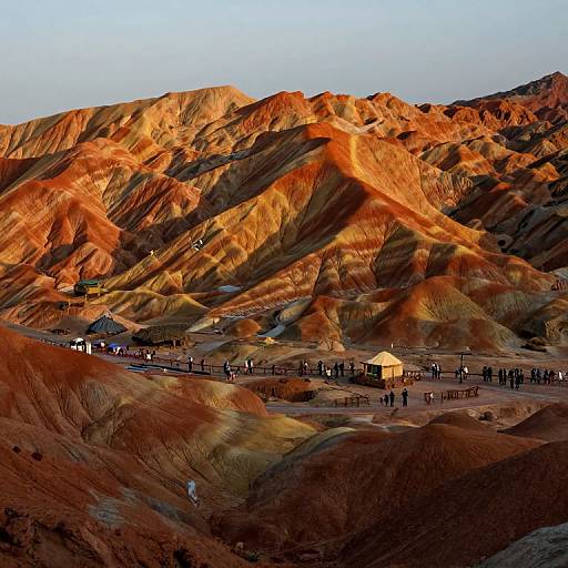 Photograph of vibrant, orange and red desert hills with winding ridges, small wooden huts, and a gathering of people in the valley under a