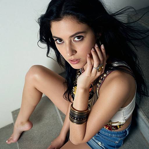 Photograph of a dark-haired woman with olive skin, sitting on gray stairs, wearing a white crop top, denim shorts, and multiple bracelets, hand