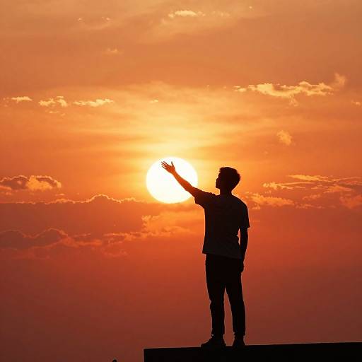 Silhouetted man stands on ledge, arm raised, against vibrant orange sunset sky with scattered clouds. Photographic image.