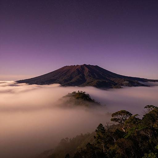 Photograph of a dark, volcanic mountain silhouetted against a purple twilight sky, partially obscured by a thick layer of glowing, white mist below