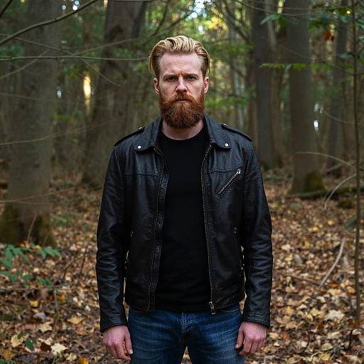 Photograph of a bearded, blond-haired man with fair skin, wearing a black jacket and jeans, standing in a forest with fallen leaves and tall