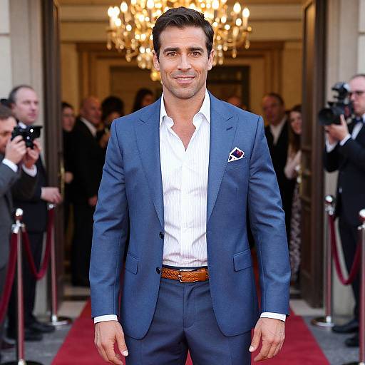 Photograph of a handsome man with short dark hair, wearing a blue suit, white shirt, and brown belt, standing on a red carpet in front