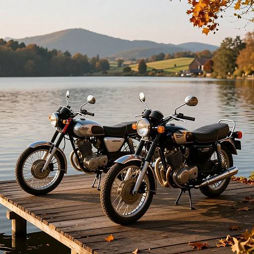 Two classic black motorcycles parked on a wooden dock by a serene lake, with autumn leaves and rolling hills in the background.
