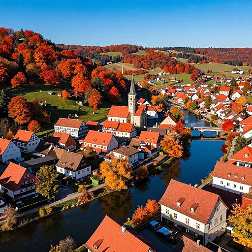 Aerial photograph of a vibrant, autumnal village with red-roofed houses, a central church, and a river, surrounded by colorful fall foliage
