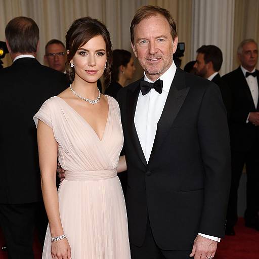 Photograph of Jennifer Lopez in a white, V-neck gown with a silver necklace, standing beside George Clooney in a black tuxedo. Background