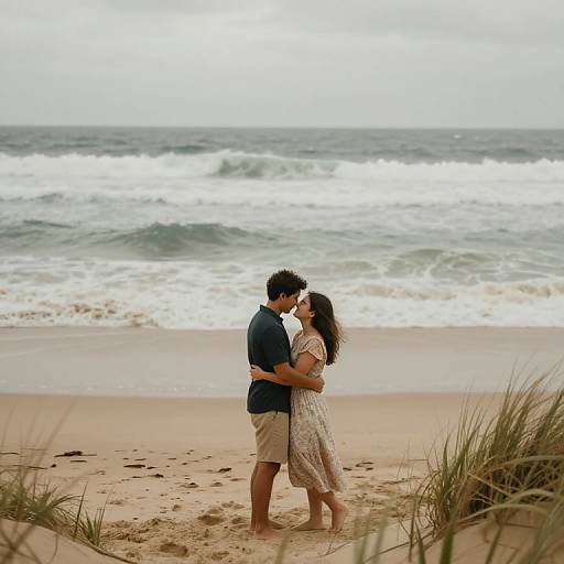 Photograph of a romantic beach scene: a couple embraces on sandy shore, waves crashing behind, overcast sky, both barefoot, casual summer attire