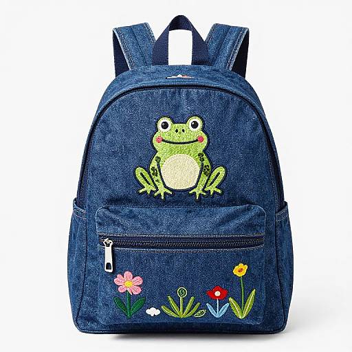 Photograph of a blue denim backpack with a green cartoon frog embroidered on the front, and colorful flowers embroidered below a zipper pocket.