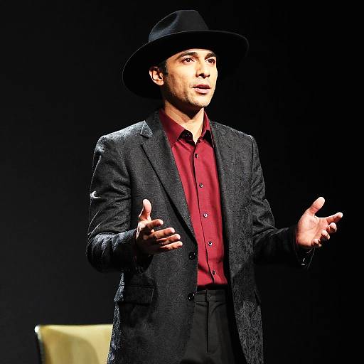 Photograph of a man with medium brown skin, wearing a black hat, red shirt, and black textured blazer, gesturing with hands on a