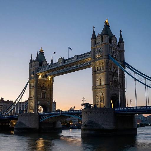 Photograph of London's Tower Bridge at dusk, with illuminated Gothic towers, blue sky, and calm river reflecting bridge lights.
