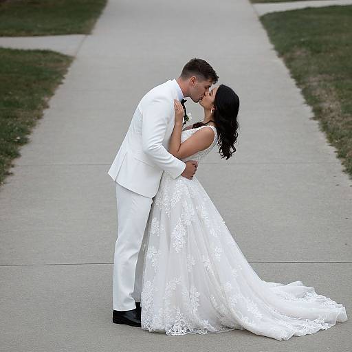 Photograph of a couple kissing on a concrete path; groom in white suit, bride in white lace wedding dress, dark hair.