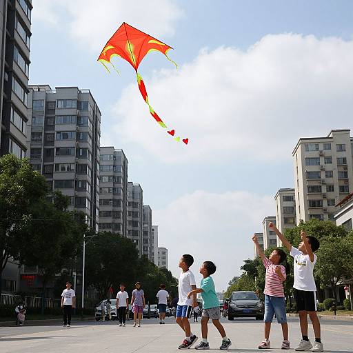 Photograph of children flying a bright red kite in an urban street with high-rise buildings, clear sky, and other kids walking.