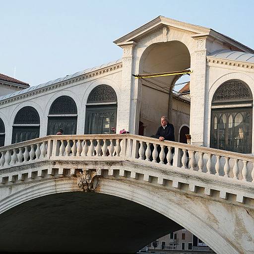 Photograph of a white, arched bridge with two people standing on the balcony of a pavilion, under a clear blue sky.