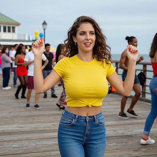 Photograph of a smiling, curvy woman with wavy brown hair, wearing a yellow crop top and blue jeans, raising hands in a lively pier