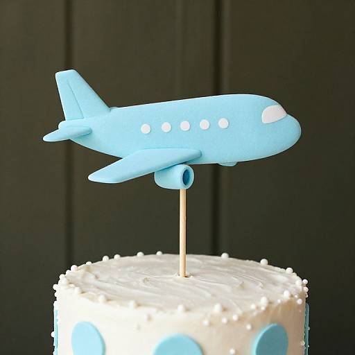 Photograph of a light blue airplane-shaped cake topper with white windows, standing on a white iced cake with blue polka dots, against a