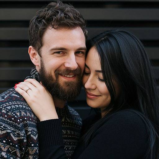 Photograph of a smiling bearded man with dark curly hair and a black patterned sweater, being affectionately hugged by a black-haired woman in a
